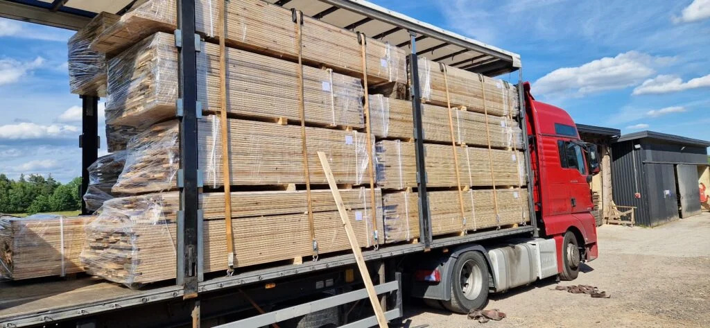 Ciężarówka z czerwonym ciągnikiem siodłowym załadowana paletami z drewnem kominkowym, zabezpieczonym folią, na tle błękitnego nieba i budynku magazynowego.