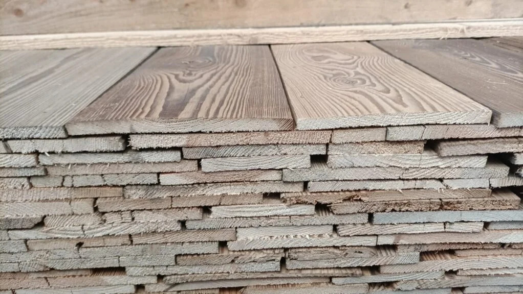Stos równo ułożonych, prostokątnych desek z widocznym usłojeniem, o różnych odcieniach brązu i szarości, przygotowanych do transportu lub składowania.