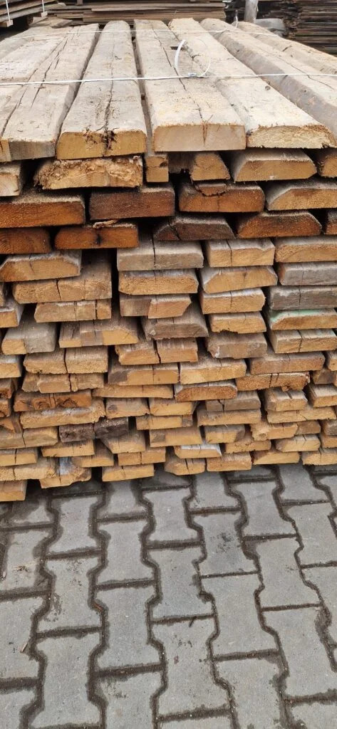 Stos równo ułożonych, szorstkich desek o nieregularnych krawędziach, zabezpieczonych białym sznurkiem, na tle szarej kostki brukowej.