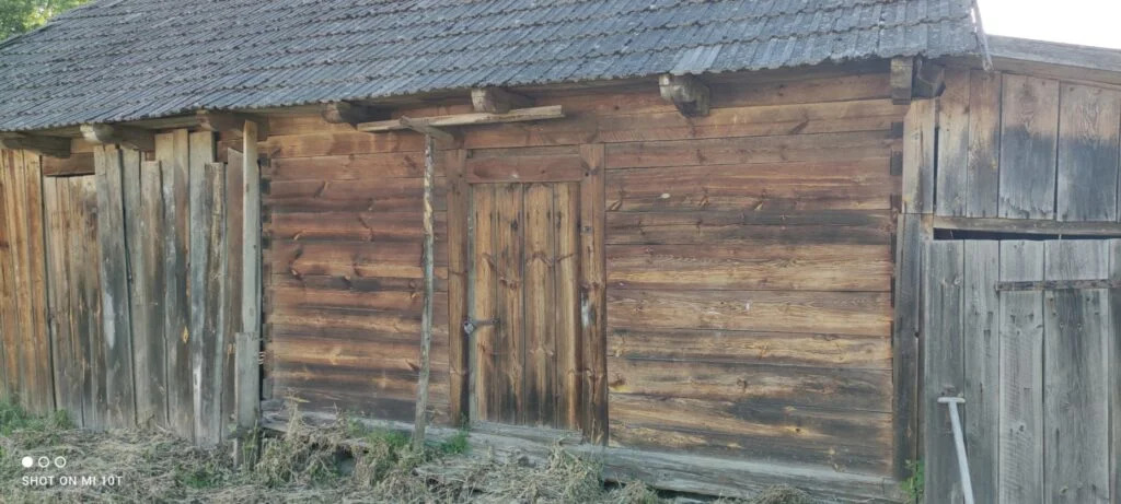 Stary, drewniany budynek gospodarczy z ciemnym, spadzistym dachem pokrytym dachówką, widoczne poziome deski z wyraźnymi słojami i drzwi z metalowym zamkiem.