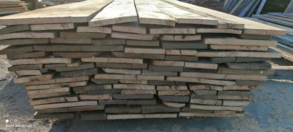 Stos ułożonych desek z widocznymi śladami cięcia, nierówne krawędzie, różne odcienie drewna, widoczne słoje i sęki.
