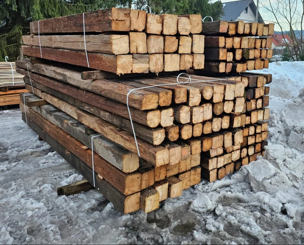 Stosy przyciętych, kwadratowych kloców drewna ułożone na śniegu, spięte białym sznurkiem, z widocznymi słojami i różnicami w odcieniach brązu.