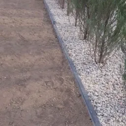 Ścieżka z ziemi obok rabaty z kamykami i iglakami, oddzielona szarym obrzeżem. Widoczne ślady opon na ziemi. Ujęcie z poziomu gruntu.