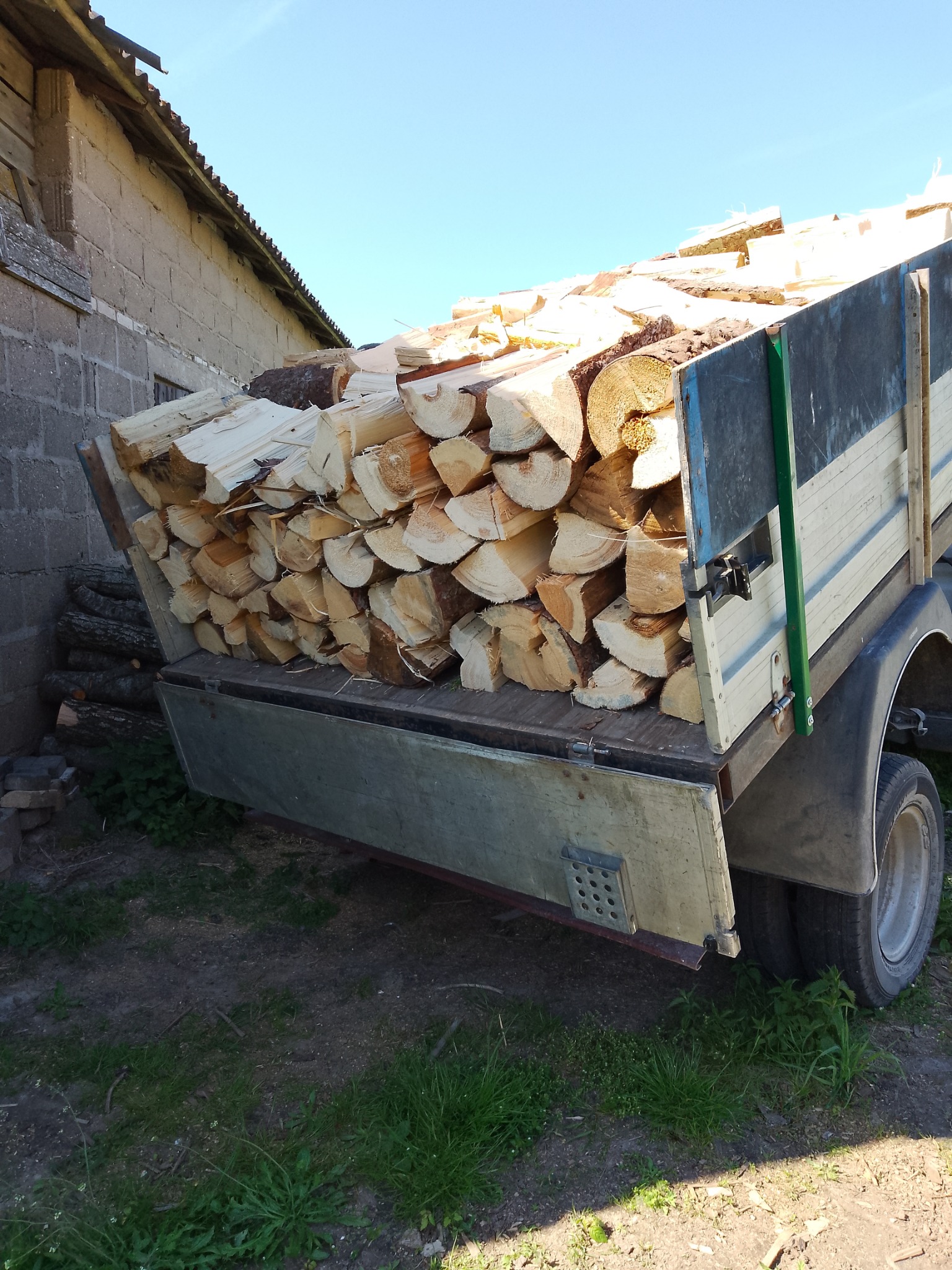 Przyczepa wypełniona pociętym drewnem kominkowym, widoczna na tle murowanego budynku i błękitnego nieba.