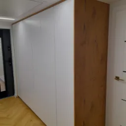 Długa szafa wnękowa z białymi frontami i wykończeniem w kolorze drewna, widoczna na tle parkietu ułożonego w jodełkę.