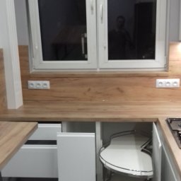 Ergo Meble - Biała kuchnia na wymiar z drewnianym blatem, widoczne szafki z systemem obrotowym i szuflady, okno nad blatem z odbiciem osoby.