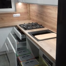 Ergo Meble - Fragment nowoczesnej kuchni z drewnianym blatem, czarną płytą grzewczą i zlewozmywakiem oraz wysuwanym systemem sortowania odpadów z kolorowymi pokrywami.