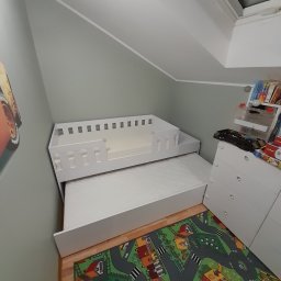 Ergo Meble - Białe, dwupoziomowe łóżko dziecięce z barierką na górnym poziomie, wysunięte z pod spodu, na podłodze kolorowy dywan z motywem miasteczka, widok z góry w pokoju na poddaszu.