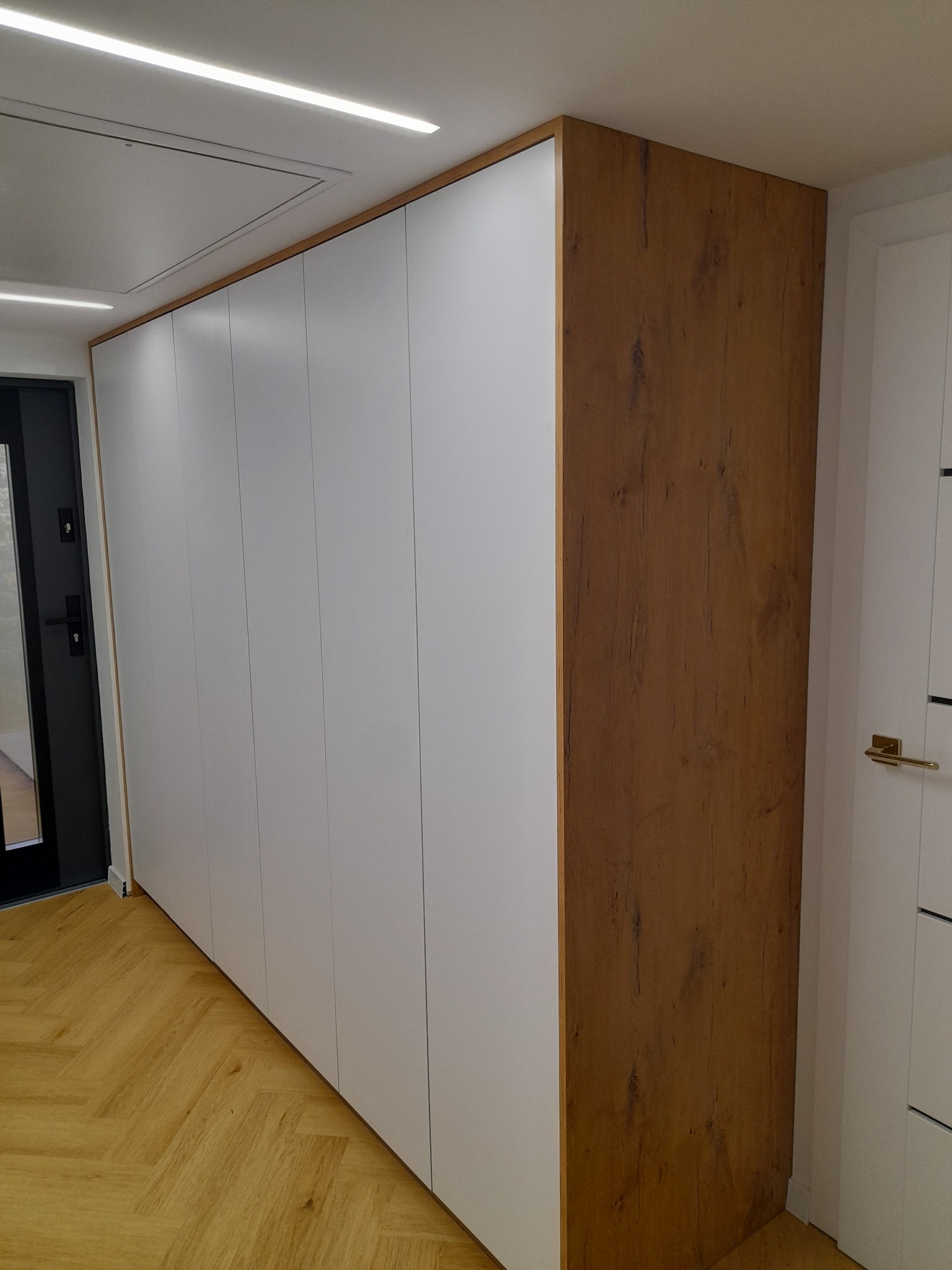 Długa szafa wnękowa z białymi frontami i wykończeniem w kolorze drewna, widoczna na tle parkietu ułożonego w jodełkę.