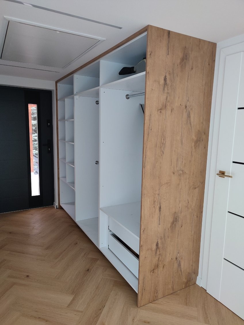 Wnęka z białą szafą z drewnopodobnymi bokami, z szufladami i półkami, widok na podłogę z parkietu i drzwi wejściowe.