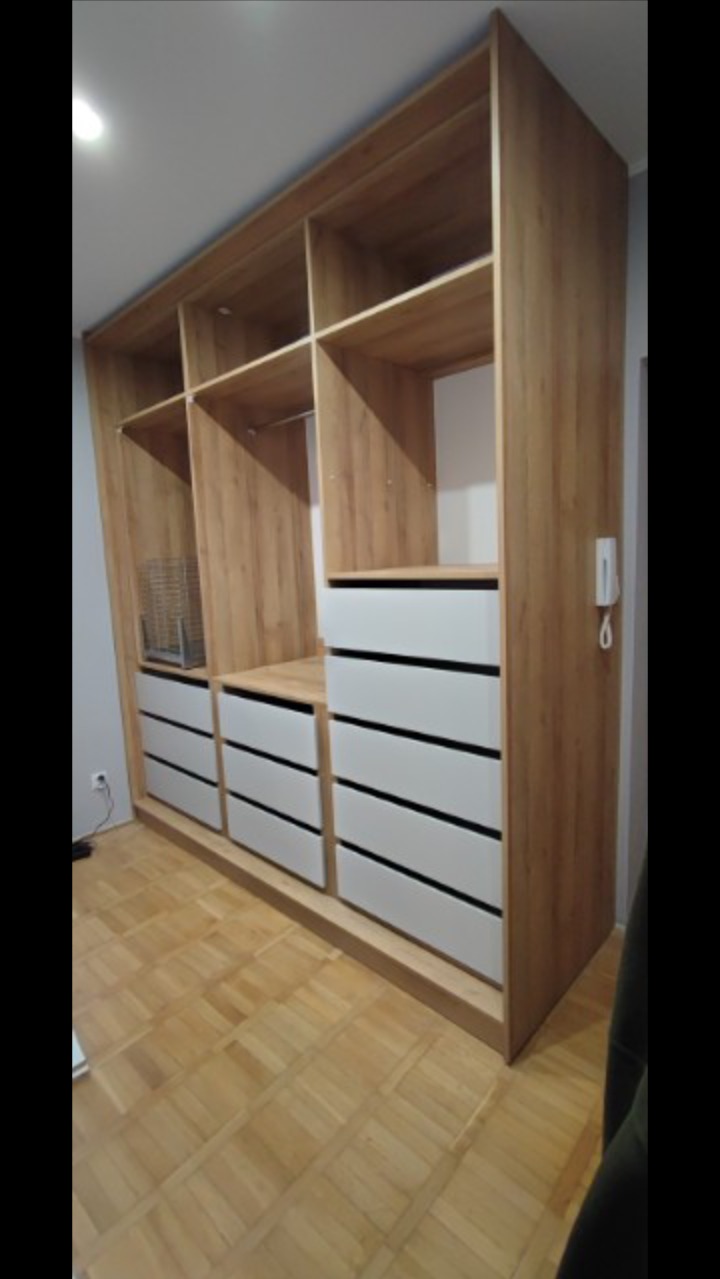 Duża, drewniana szafa z białymi szufladami, wbudowana w ścianę, z widoczną parkietową podłogą.