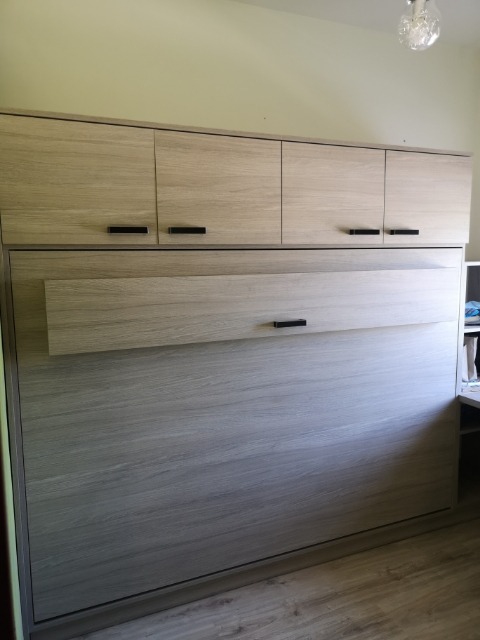 Jasna, drewnopodobna szafa wnękowa z górnymi szafkami, minimalistyczne czarne uchwyty, widoczna podłoga i lampa sufitowa.