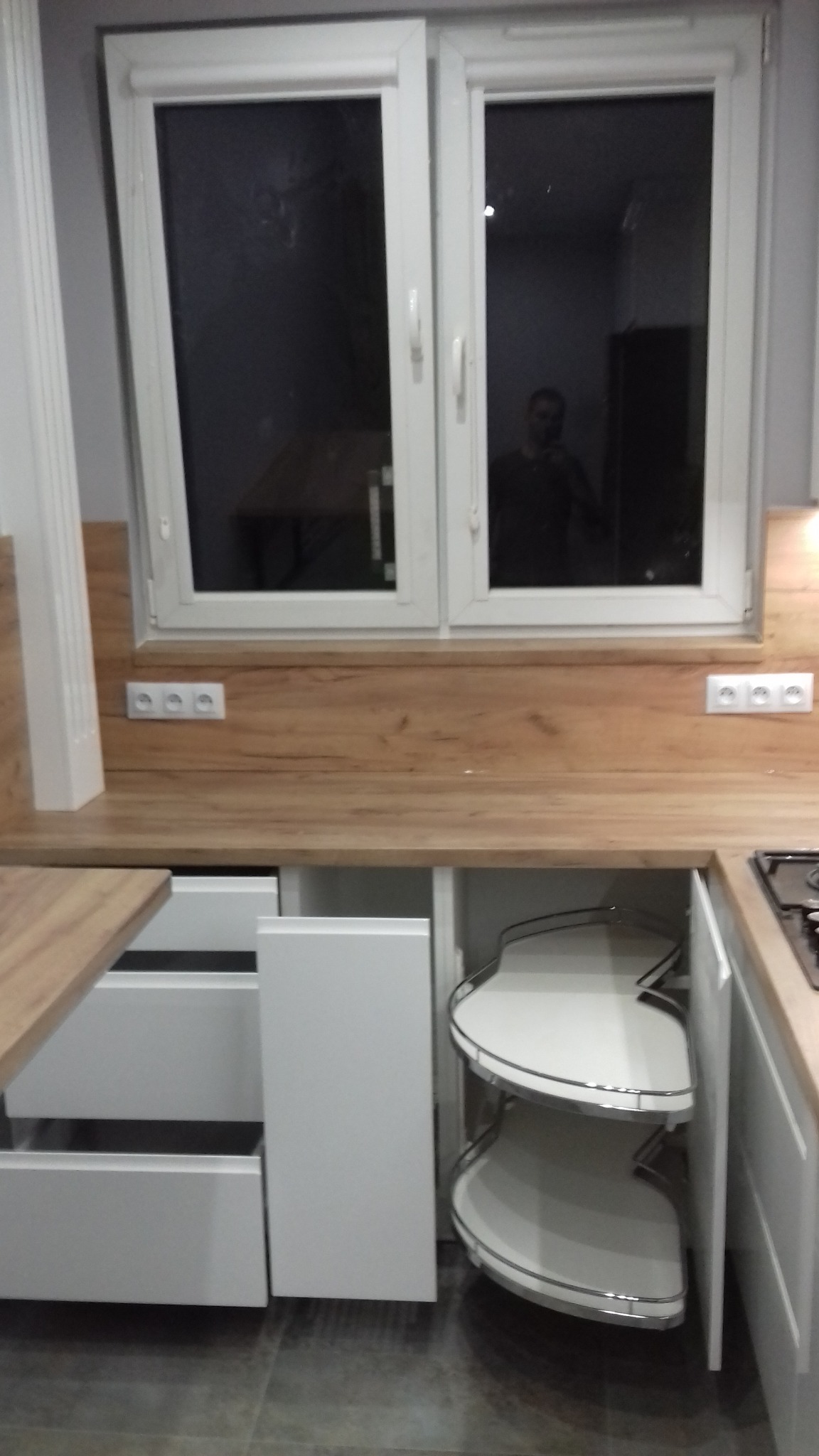 Biała kuchnia na wymiar z drewnianym blatem, widoczne szafki z systemem obrotowym i szuflady, okno nad blatem z odbiciem osoby.