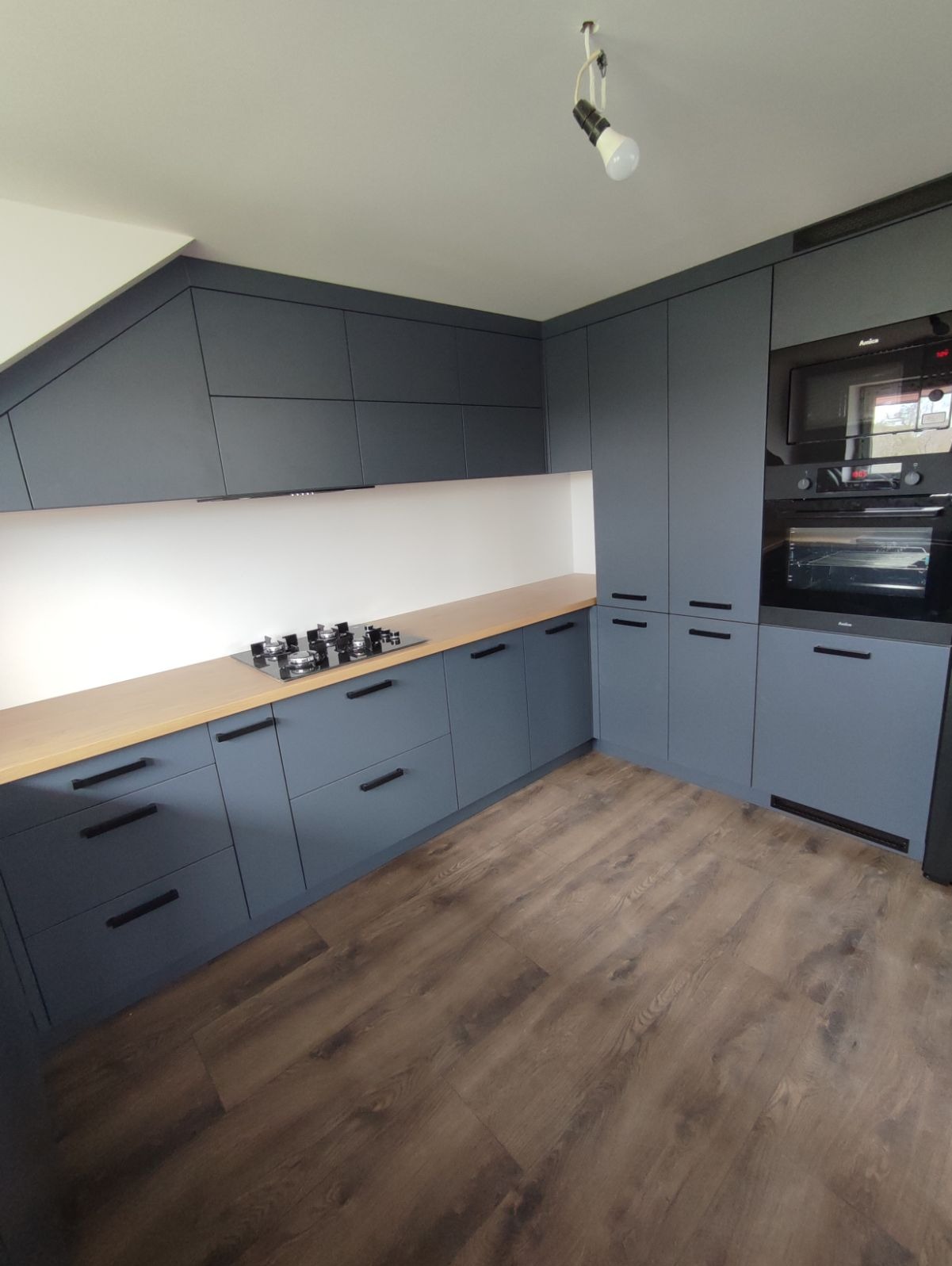 Nowoczesna, szara kuchnia z drewnianym blatem i czarnymi uchwytami, oświetlona punktowym światłem. Zabudowa pod skosem, płyta gazowa na blacie, piekarnik i mikrofalówka w zabudowie.