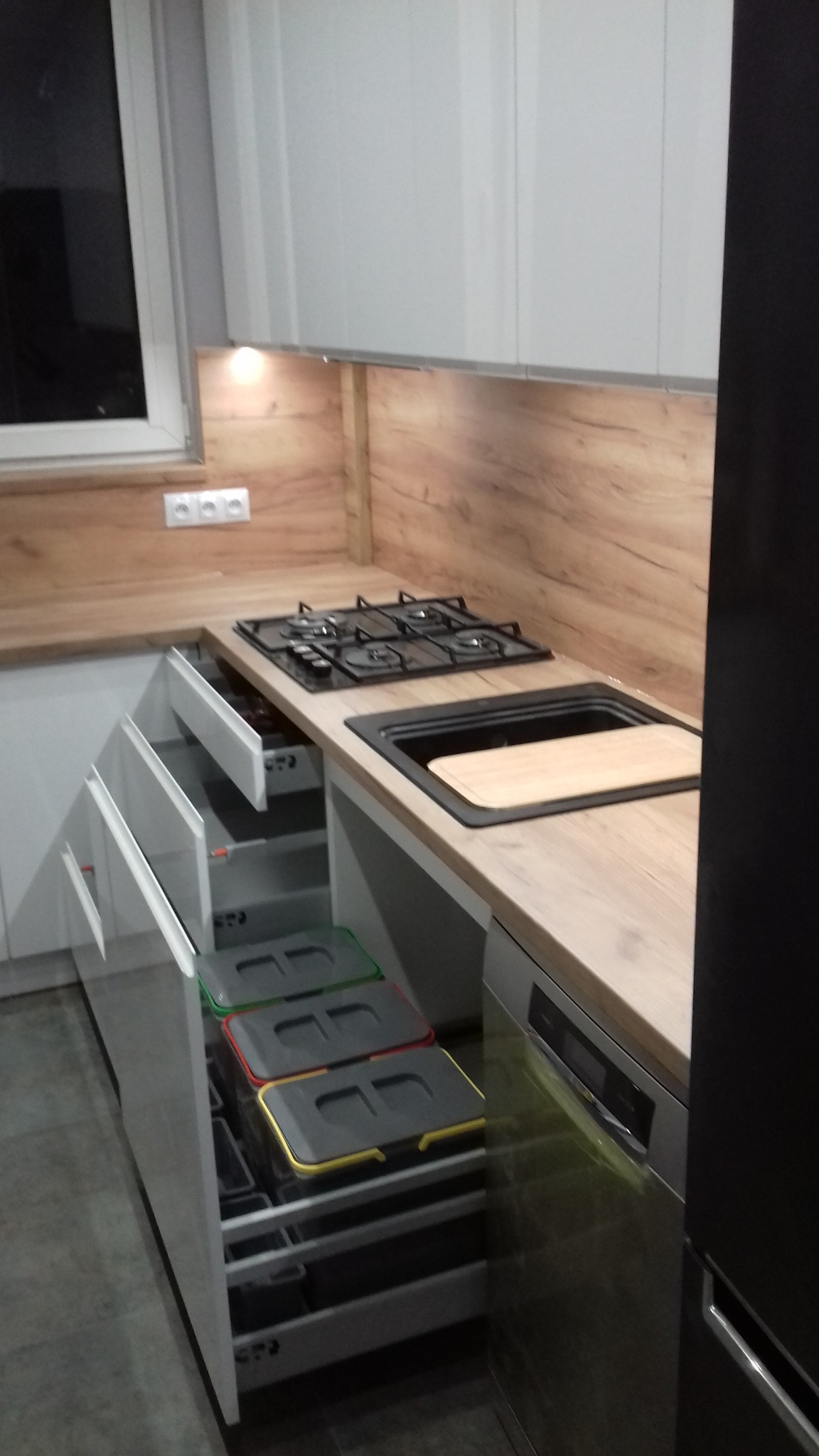 Fragment nowoczesnej kuchni z drewnianym blatem, czarną płytą grzewczą i zlewozmywakiem oraz wysuwanym systemem sortowania odpadów z kolorowymi pokrywami.