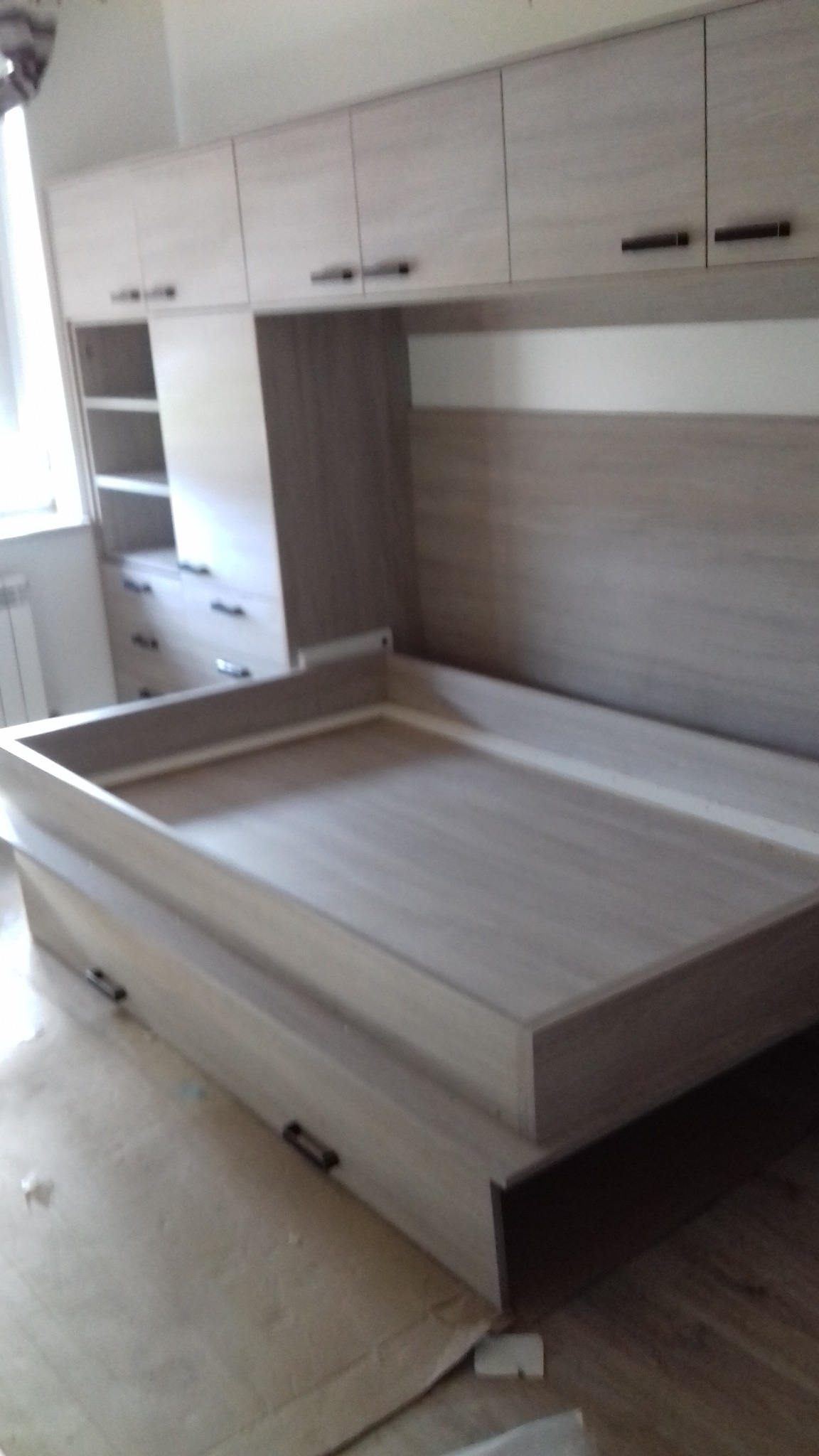 Beżowe meble do sypialni z szafkami wiszącymi i łóżkiem ze schowkami, widoczne szuflady i półki, minimalistyczny styl, oświetlenie dzienne.