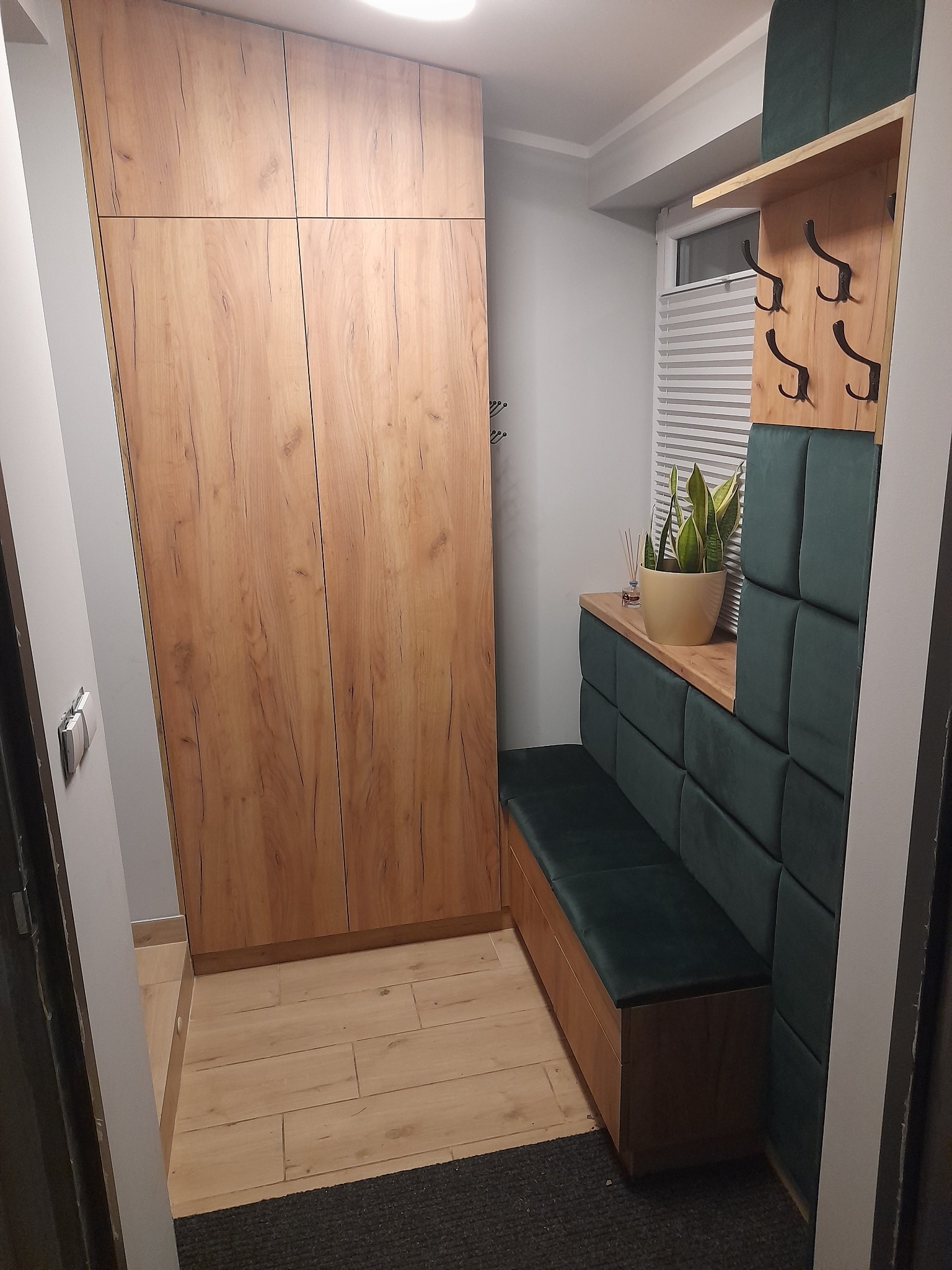 Przedpokój z zabudową: wysoka szafa z drewnopodobnymi frontami, siedzisko z zielonym tapicerowanym panelem ściennym, drewniana półka z wieszakami.