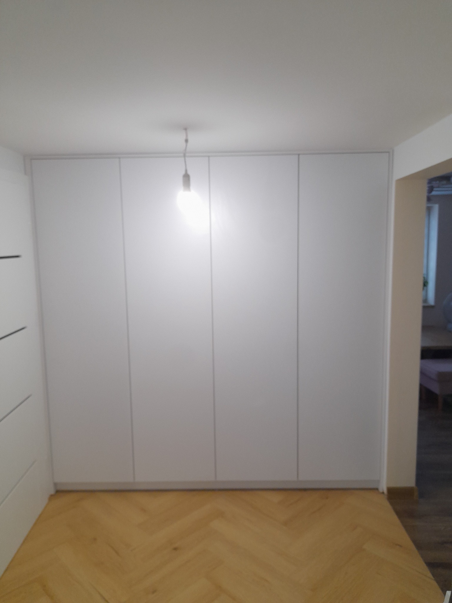 Biała, minimalistyczna szafa wnękowa z czterema frontami, zamontowana w przedpokoju z parkietem w jodełkę.