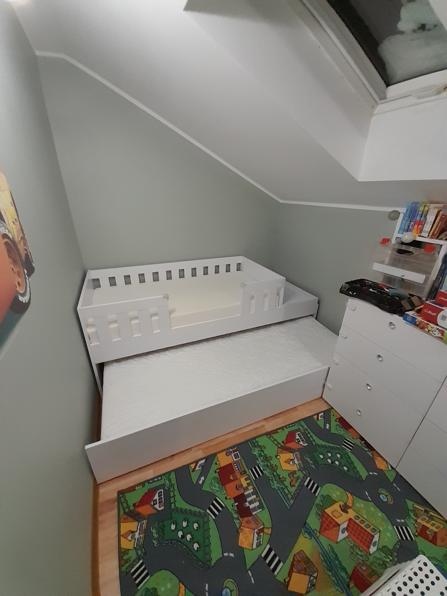 Białe, dwupoziomowe łóżko dziecięce z barierką na górnym poziomie, wysunięte z pod spodu, na podłodze kolorowy dywan z motywem miasteczka, widok z góry w pokoju na poddaszu.