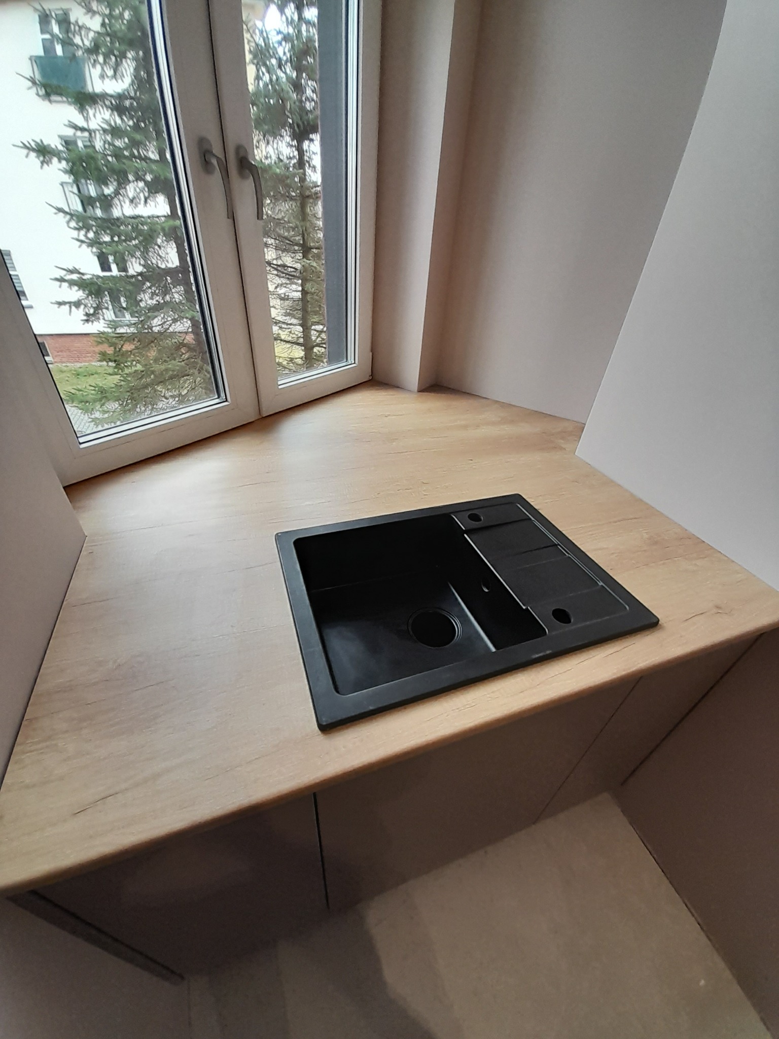 Czarny, minimalistyczny zlew z ociekaczem wkomponowany w drewniany blat pod oknem, widok z góry.