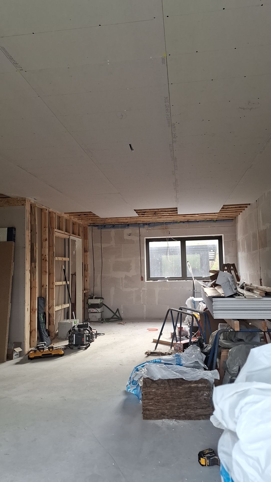 Wnętrze w trakcie montażu płyt kartonowo-gipsowych na suficie, widoczne profile drewniane, instalacja elektryczna, narzędzia i materiały budowlane w surowym stanie.