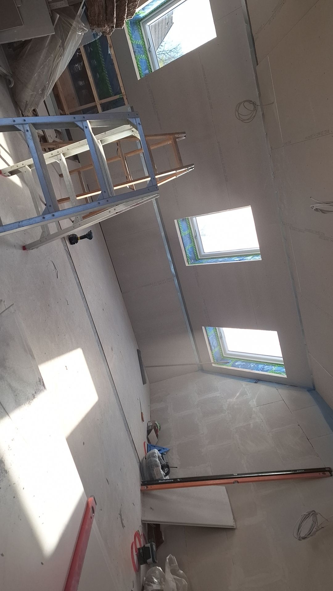 Wnętrze w trakcie montażu płyt kartonowo-gipsowych na ścianach i suficie, widoczne trzy okna dachowe, drabina aluminiowa oraz narzędzia budowlane.