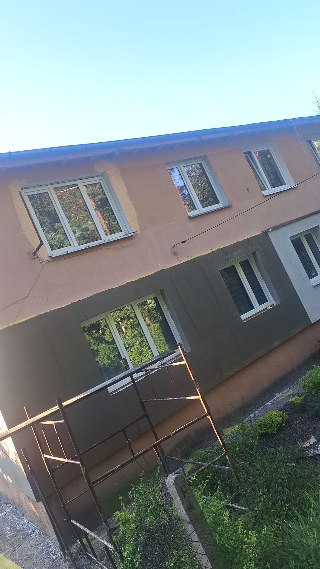 Fragment elewacji budynku w trakcie prac remontowych: część ściany ocieplona i pokryta szarą warstwą, widoczne okna z odbiciem drzew, rusztowanie w dolnej części kadru.