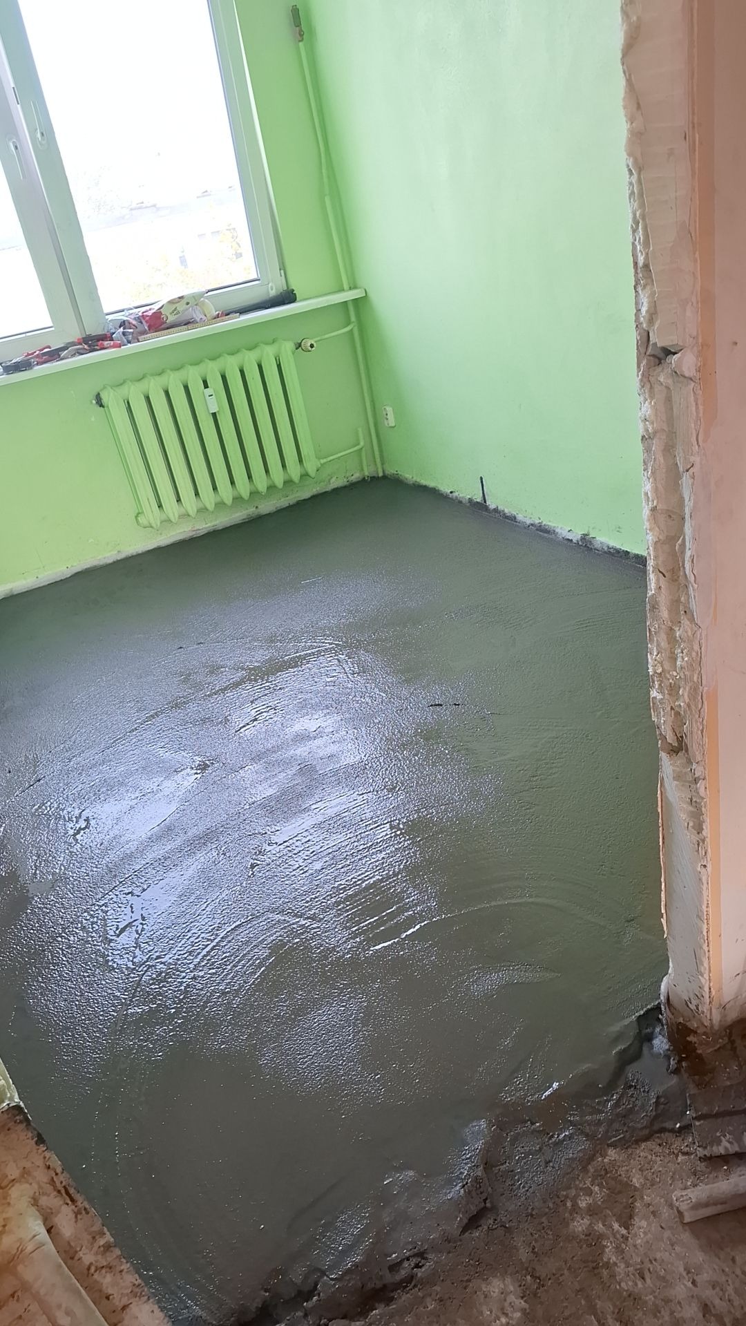 Wylany, świeży podkład podłogowy w pomieszczeniu z zielonymi ścianami i grzejnikiem, widoczne ślady po rozprowadzaniu masy.