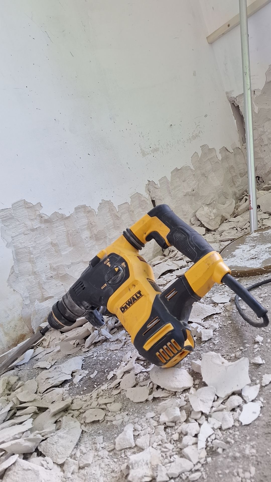 Żółto-czarny młot udarowy DeWalt leży na podłodze pokrytej odłamkami tynku, częściowo usuniętego ze ściany w tle.