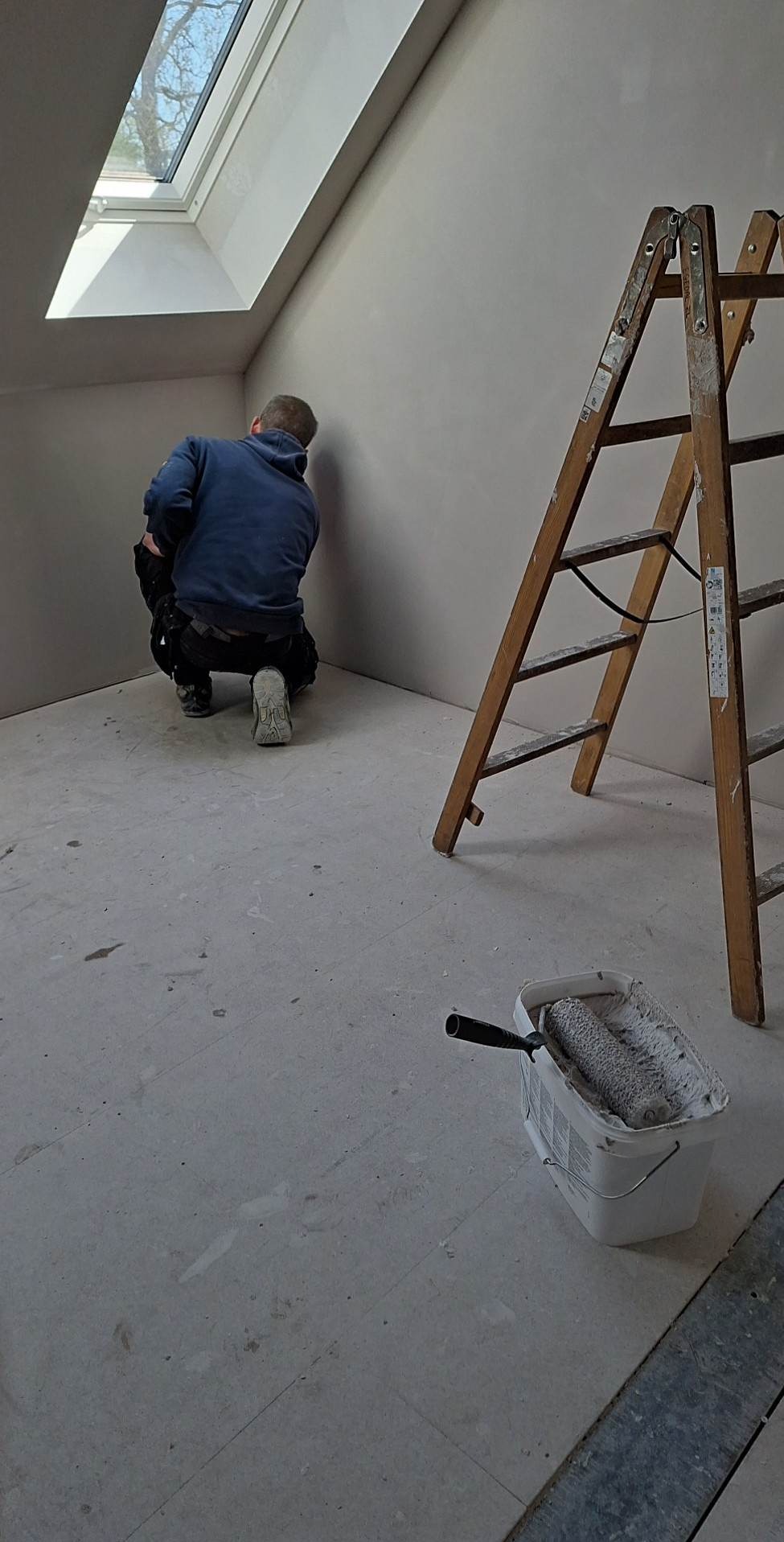 Pracownik w kucki wygładza ścianę w rogu pomieszczenia z oknem dachowym, obok stoi drewniana drabina i wiadro z wałkiem malarskim.