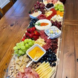 SALAMO ALIKOM SALEM ELMAADALAWI - Bogata deska serów, wędlin, owoców i słodyczy z akcentem wielkanocnym (jajka czekoladowe), ułożona na drewnianym stole. Widoczne sery z dekoracją marchewki, winogrona, truskawki, orzechy, szynka...