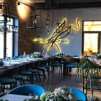 Uroczysta aranżacja sali restauracyjnej z długimi stołami udekorowanymi kwiatami, turkusowymi krzesłami i oświetlonym logo firmy na ścianie z cegły.