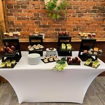 Stół przykryty białym obrusem, na którym znajdują się różnorodne desery i ciasta na czarnych, wielopoziomowych paterach. W tle widoczna ściana z czerwonej cegły i rośliny doniczkowe.