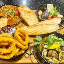 Zestaw owoców morza i kalmarów w trzech czarnych miseczkach, podany z pieczywem na drewnianej desce. Kalmary w panierce, kalmary w sosie pomidorowym i owoce morza z cytryną i ziołami.