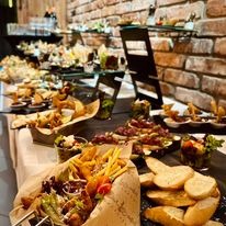 Stół cateringowy z różnorodnymi przekąskami: smażone frytki w papierowej wytłoczce, kanapeczki, sałatki w szklanych naczyniach, ustawione na białym obrusie z ceglanym tłem.