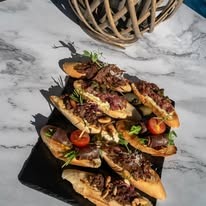 Kanapki na bagietkach z ciemnym farszem, pomidorkami cherry i ziołami, ułożone na czarnym talerzu na marmurowym blacie, z dekoracją z wikliny w tle.