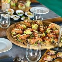 Elegancko podane przekąski na drewnianej tacy, w tle szklane naczynia z sałatkami i kieliszki, całość na szarym obrusie z elementami dekoracyjnymi w odcieniach zieleni.