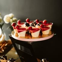 Deserowe pucharki z białą bazą i czerwoną galaretką zwieńczone świeżymi malinami, ustawione na złotej tacy z różową obwódką, na czarnym, błyszczącym postumencie.