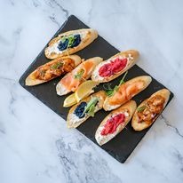 Zestaw eleganckich kanapek na ciemnej, kamiennej desce. Różne rodzaje: z kawiorem, łososiem, pastą z czerwonych buraków i krewetkami. Dekoracja z cytryny i rukoli.