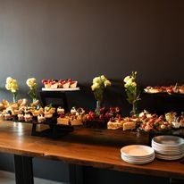 Bogato zastawiony stół z różnorodnymi ciastami, deserami i przekąskami cateringowymi, udekorowany bukietami białych i czerwonych kwiatów w szklanych wazonach, na tle ciemnej ściany, z ułożonymi...