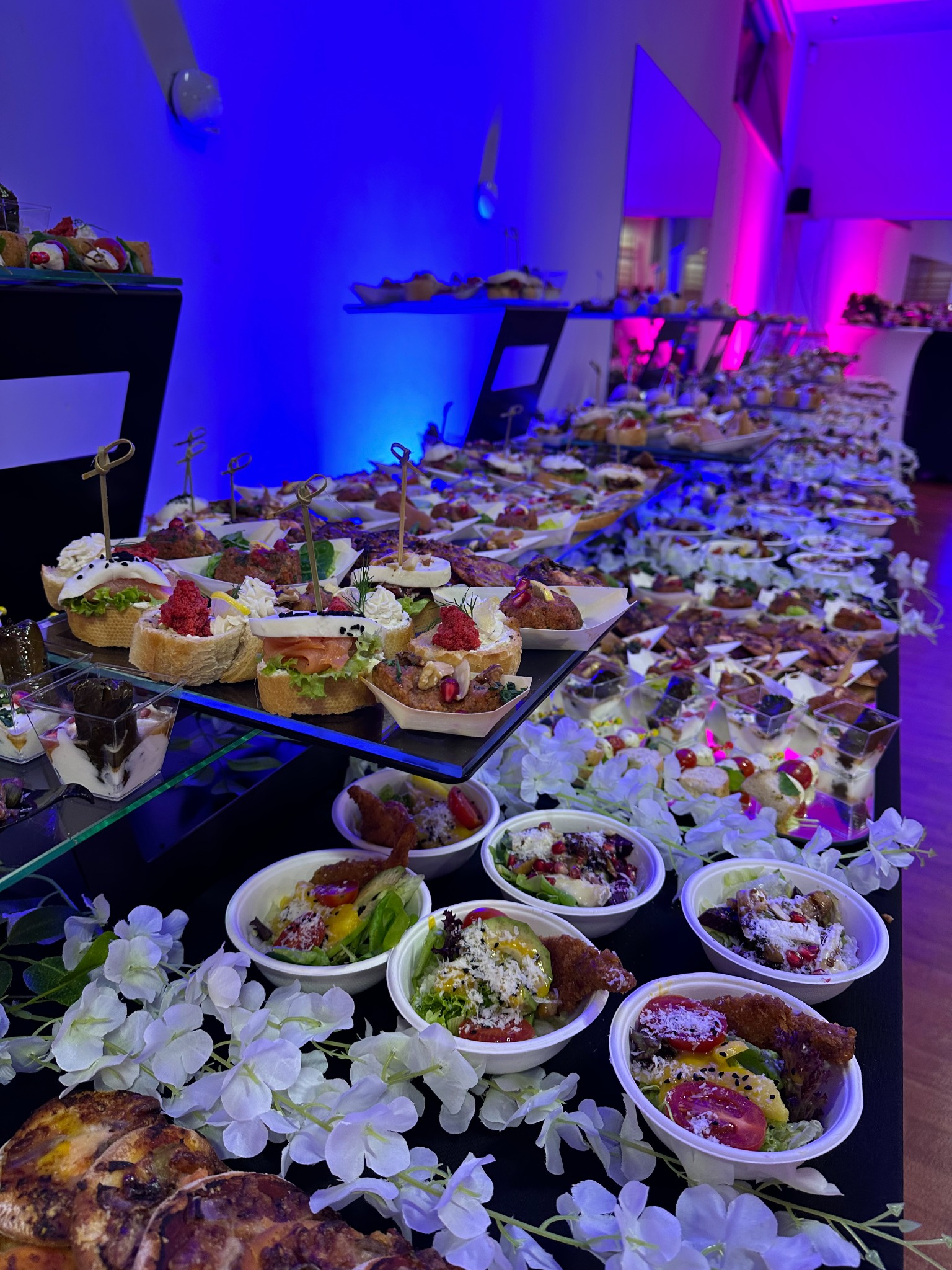 Długi stół bankietowy zastawiony różnorodnymi przekąskami, w tym mini-kanapeczkami, sałatkami w małych miseczkach i deserami w szklanych naczyniach, udekorowany białymi kwiatami, oświetlony...