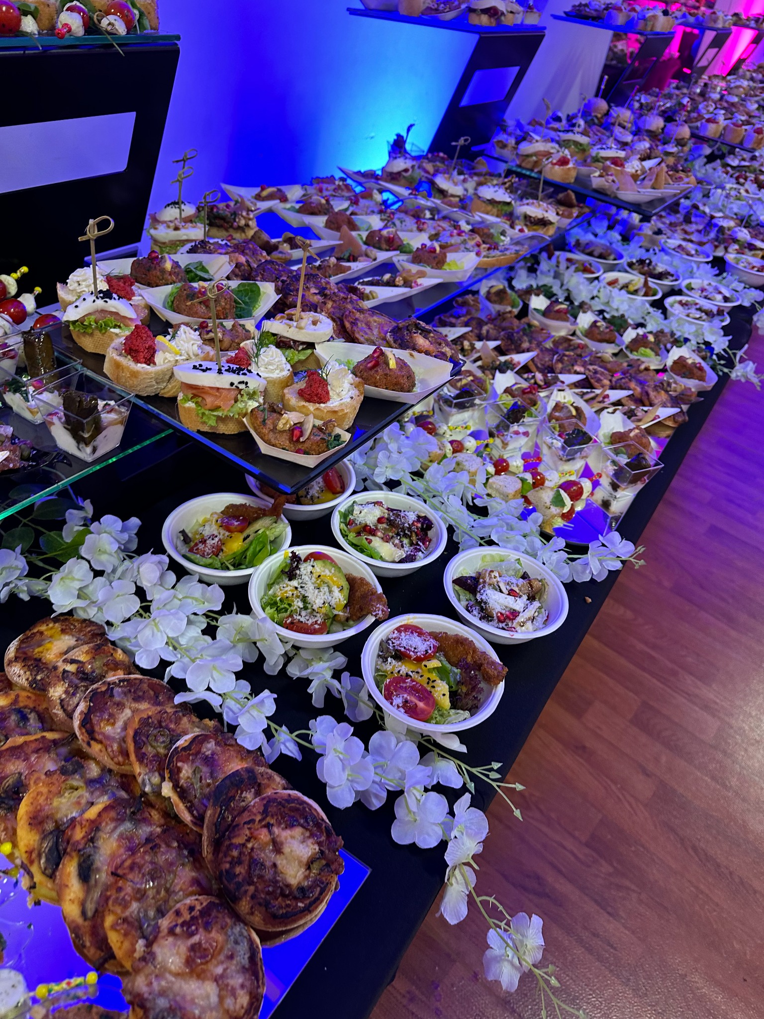 Bogato zastawiony stół z różnorodnymi przekąskami, sałatkami w miseczkach i mini kanapeczkami na szklanych paterach, udekorowany białymi kwiatami, oświetlony niebieskim światłem.