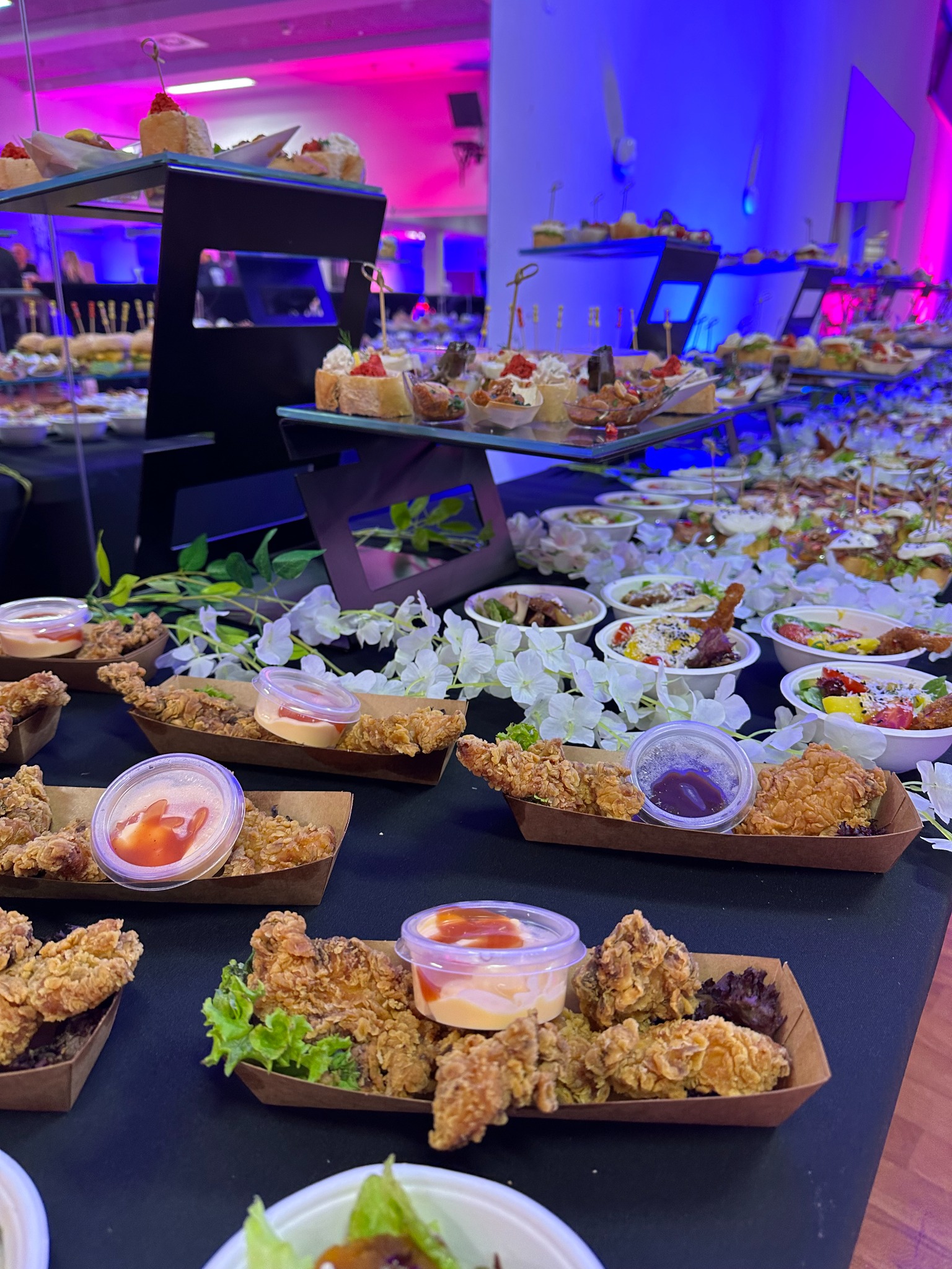 Bufet z przekąskami: smażone kurczaki w papierowych tackach z sosem, sałatki w miseczkach, kanapeczki na stojakach, dekoracje z białych kwiatów, oświetlenie w kolorach różu i błękitu.