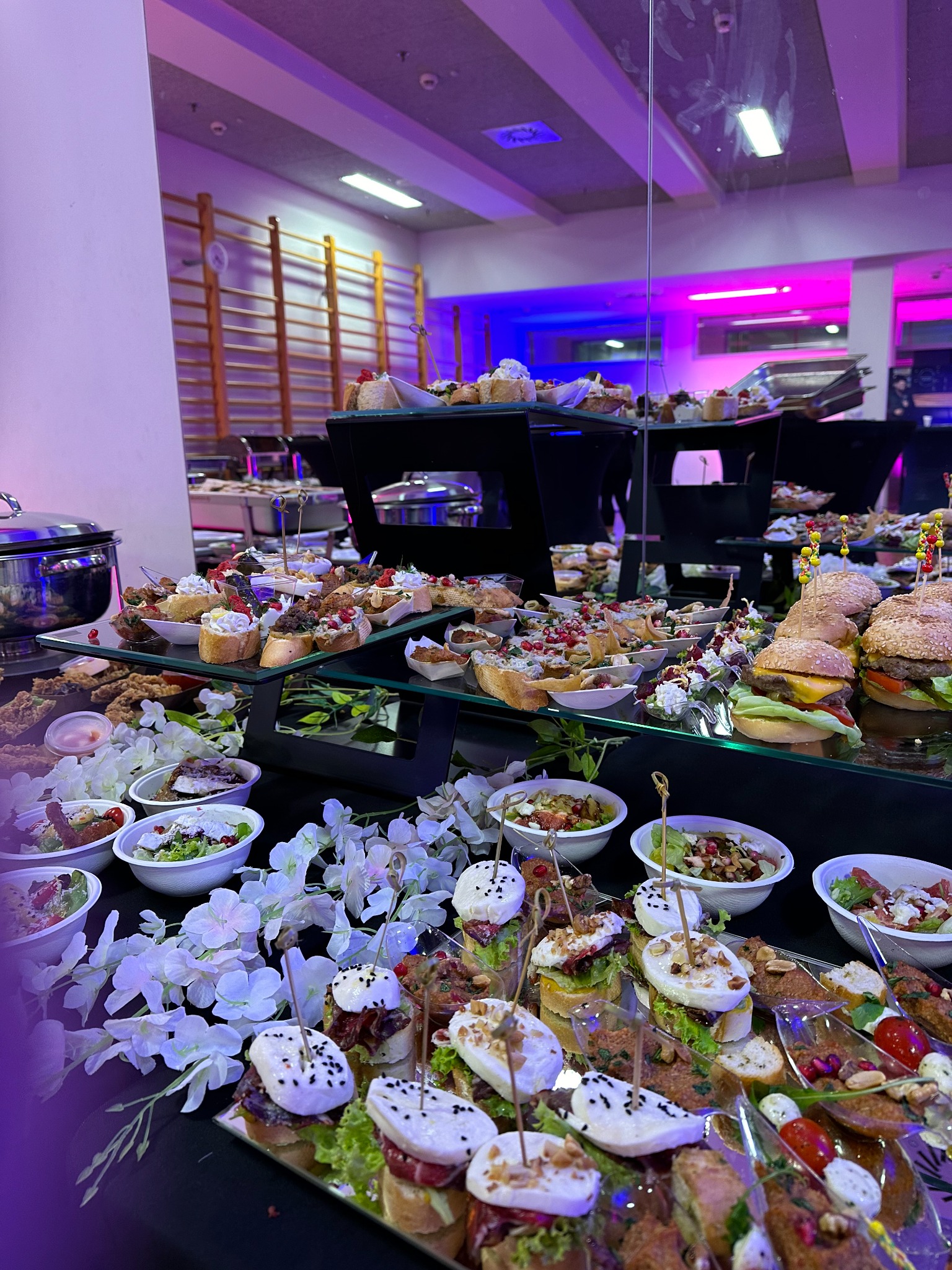 Bogato zastawiony stół z różnorodnymi przekąskami, w tym mini-burgery, kanapeczki i sałatki w małych miseczkach, udekorowany białymi kwiatami, oświetlony fioletowym światłem, z drabinkami...