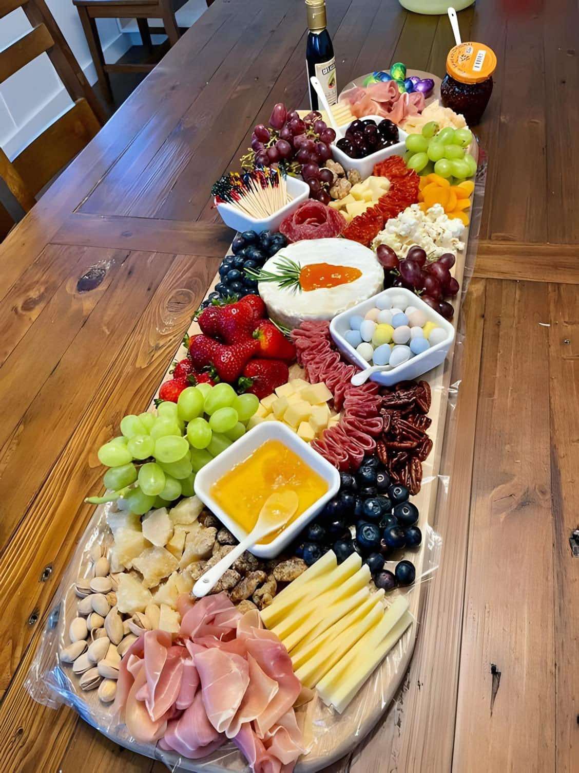 Bogata deska serów, wędlin, owoców i słodyczy z akcentem wielkanocnym (jajka czekoladowe), ułożona na drewnianym stole. Widoczne sery z dekoracją marchewki, winogrona, truskawki, orzechy, szynka...