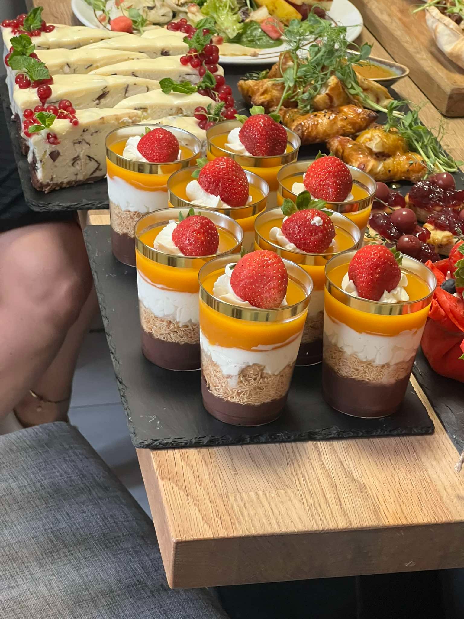 Deserowe pucharki z warstwami czekolady, kremu, mango i truskawką na wierzchu, obok sernik z porzeczkami i inne przekąski na drewnianym stole.