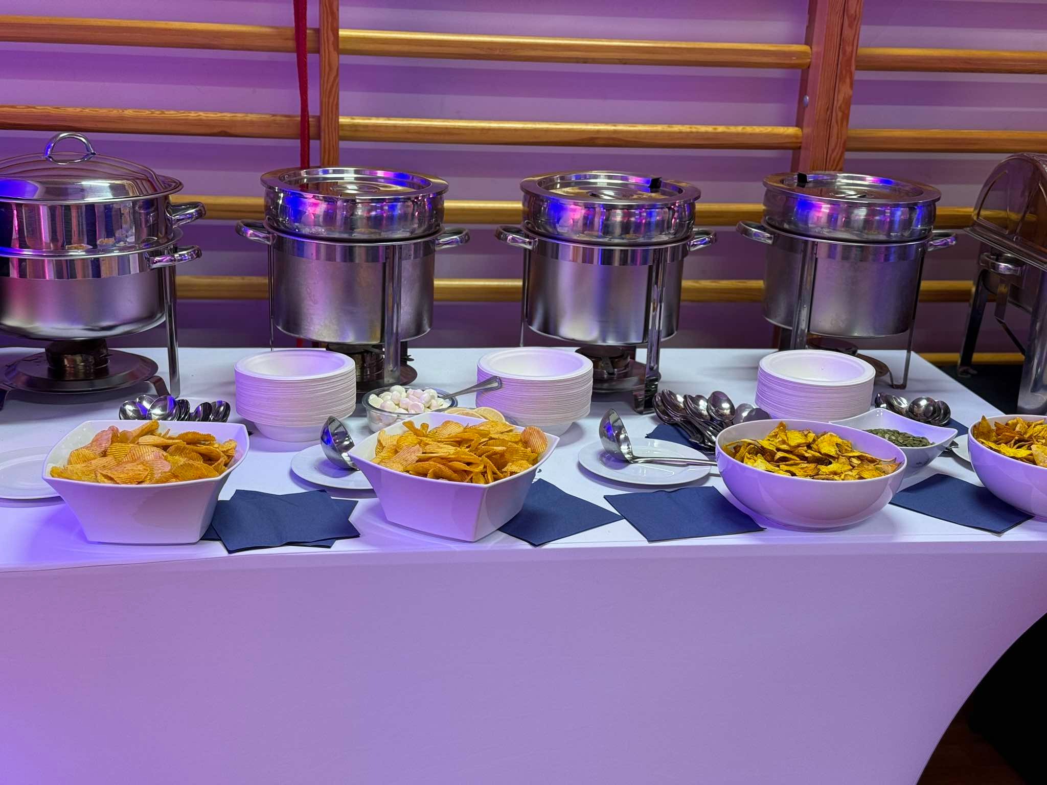 Stół cateringowy z podgrzewaczami, miskami z chipsami i przekąskami, ułożony na białym obrusie, oświetlony fioletowym światłem, z widocznymi elementami drewnianej konstrukcji w tle.