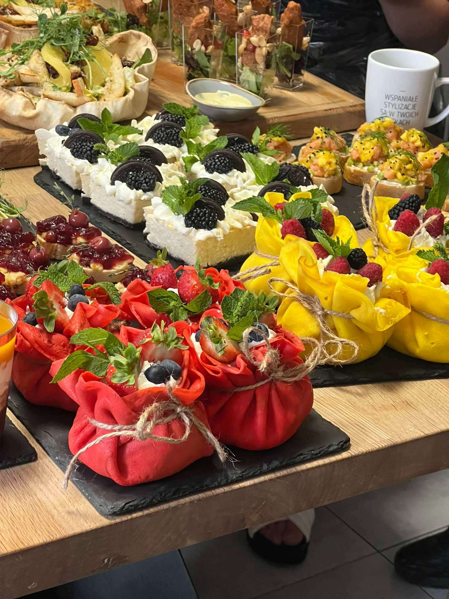 Różnorodny wybór deserów na stole, w tym tartaletki z wiśniami, sernik z Oreo, kanapki z łososiem i mango oraz naleśniki w kształcie sakiewek z owocami, wszystko na czarnych łupkowych talerzach.