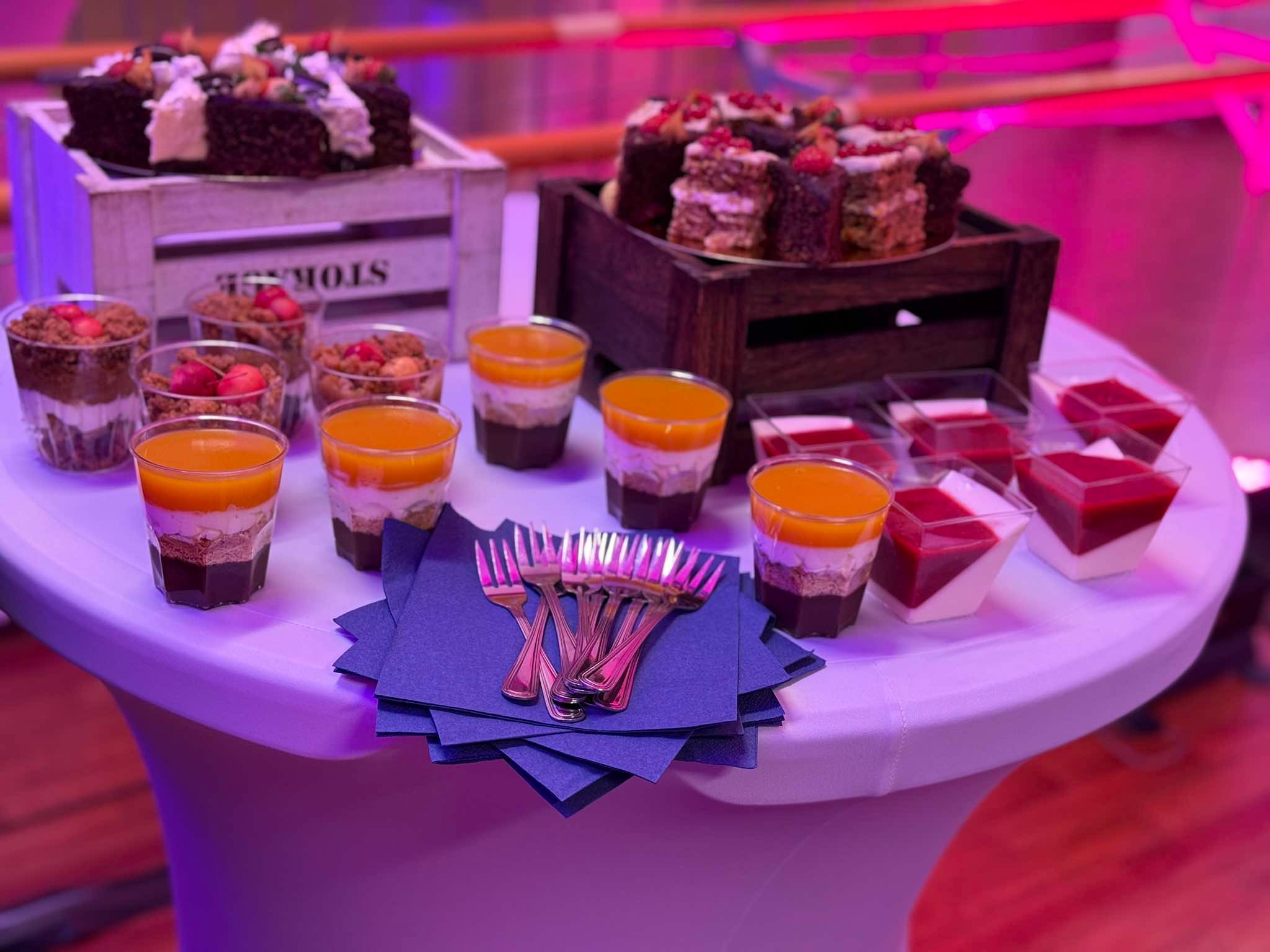 Elegancko zaaranżowany stół z deserami: mini ciasta na drewnianych podstawkach, desery warstwowe w szklankach i plastikowych pojemnikach, srebrne widelczyki na granatowych serwetkach, oświetlenie...