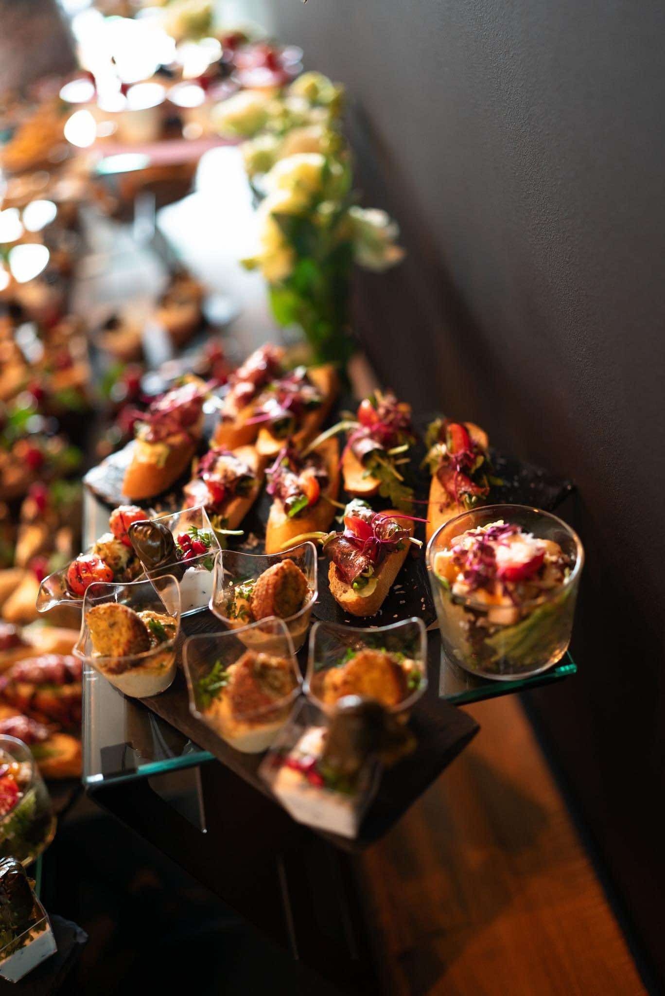 Elegancko zaaranżowane przekąski na paterach i w przezroczystych naczyniach, w tym bruschetta z pomidorami i rukolą oraz mini porcje sałatek, ustawione na szklanym stole z ciemnym tłem.